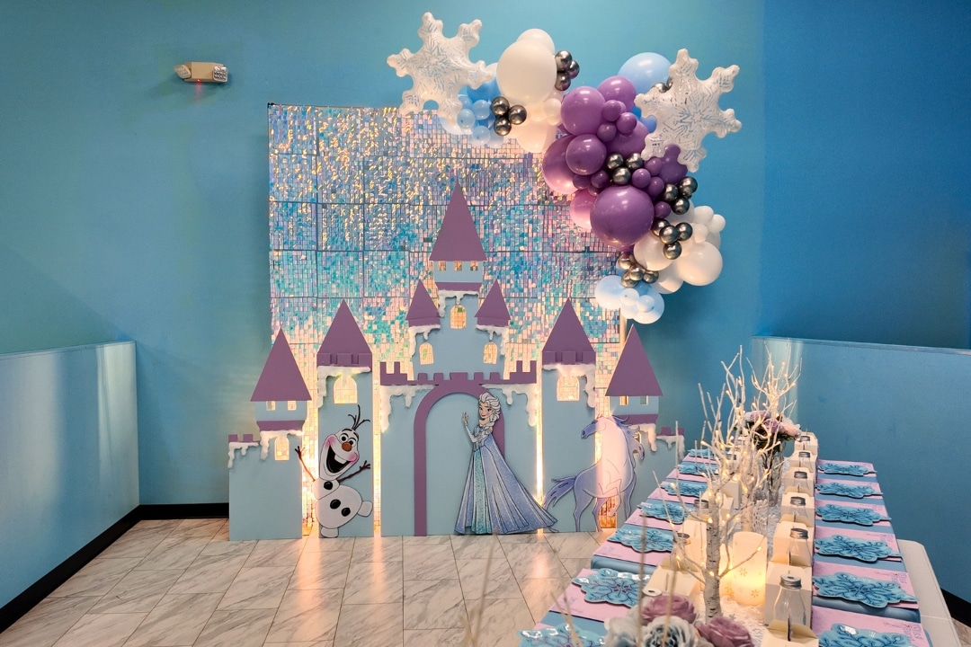 Frozen themed birthday party with illuminated ice castle backdrop, Elsa and Olaf cutouts, snowflake decor, and purple balloon garland at Kidz Kastle in Rhode Island.