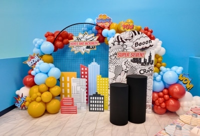 Colorful comic book and superhero cityscape backdrop for a private kids birthday party Warwick RI, showing professional balloon arches and custom party props.